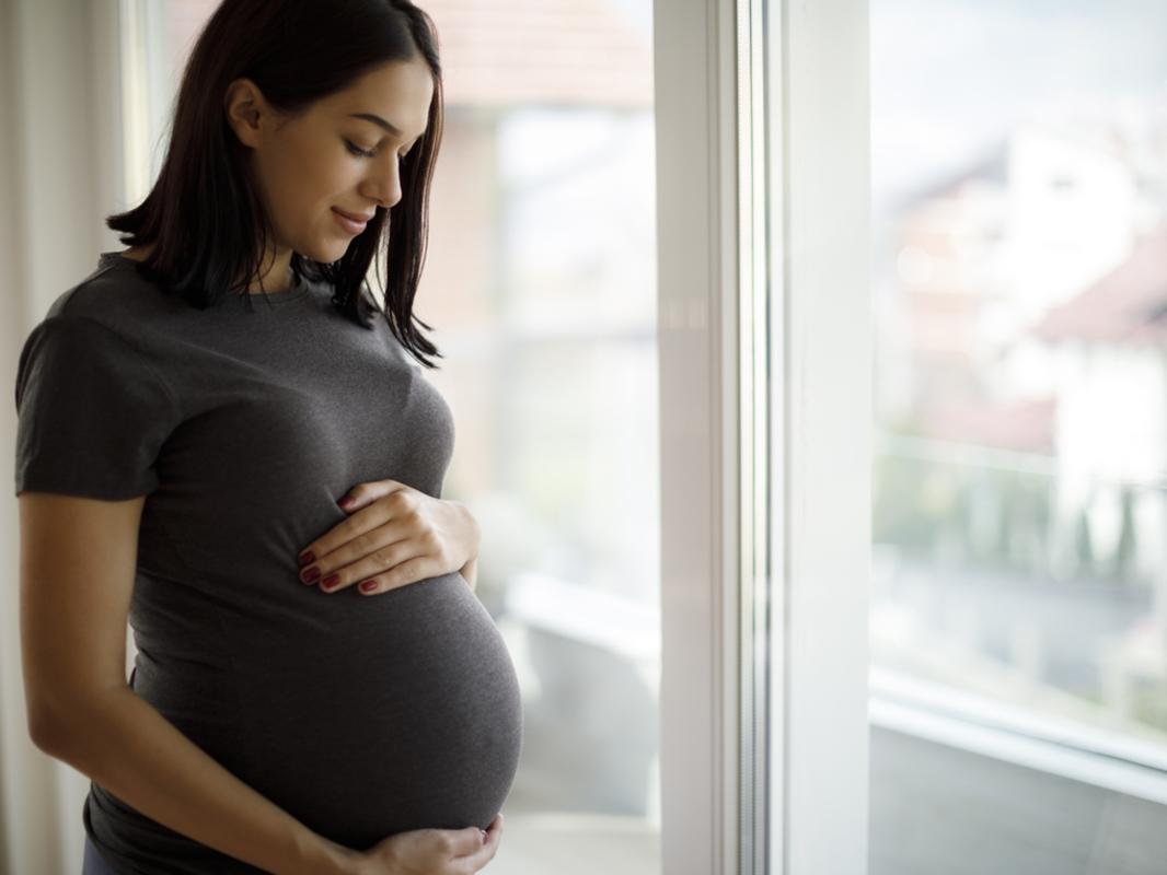A pregnant young woman with shoulder-length hair puts her hands on her belly. She is standing next to a window.