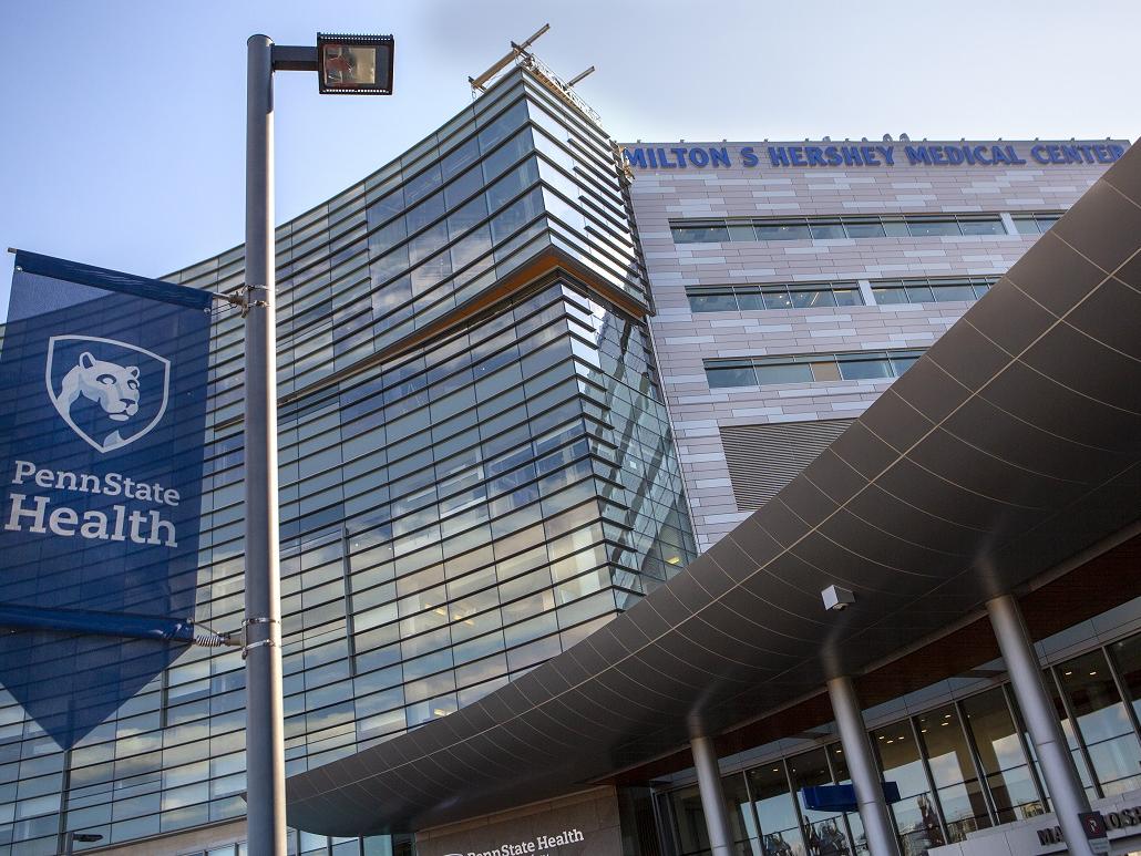 A blue and white flag hangs from a light pole in front of the entrance to Hershey Medical Center. On the flag is the Nittany Lion shield and the words, Penn State Health.
