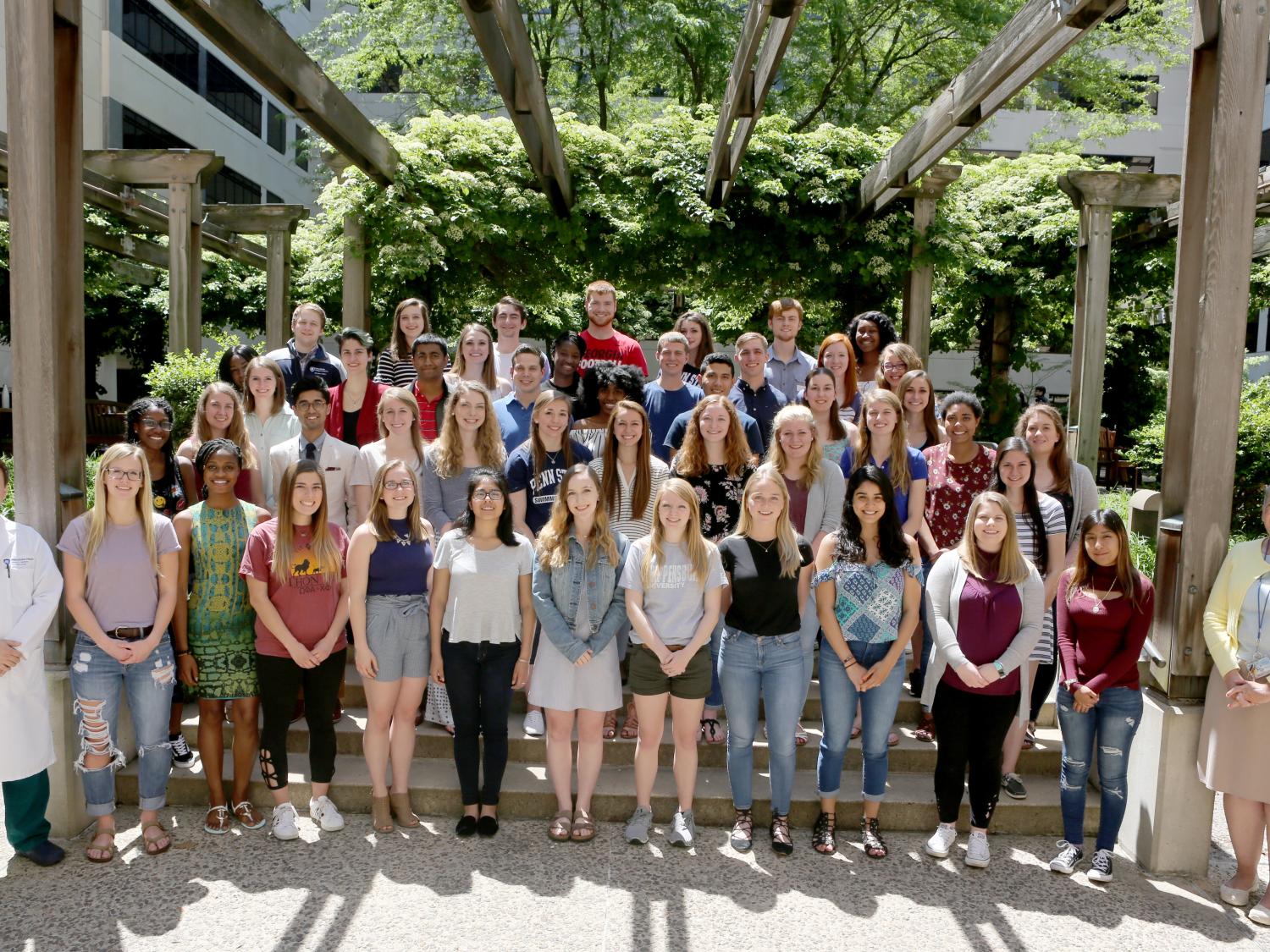 A few dozen young people pose for a photo in four rows. Green bushes and trees, as well as a white building with dark windows are in the background.