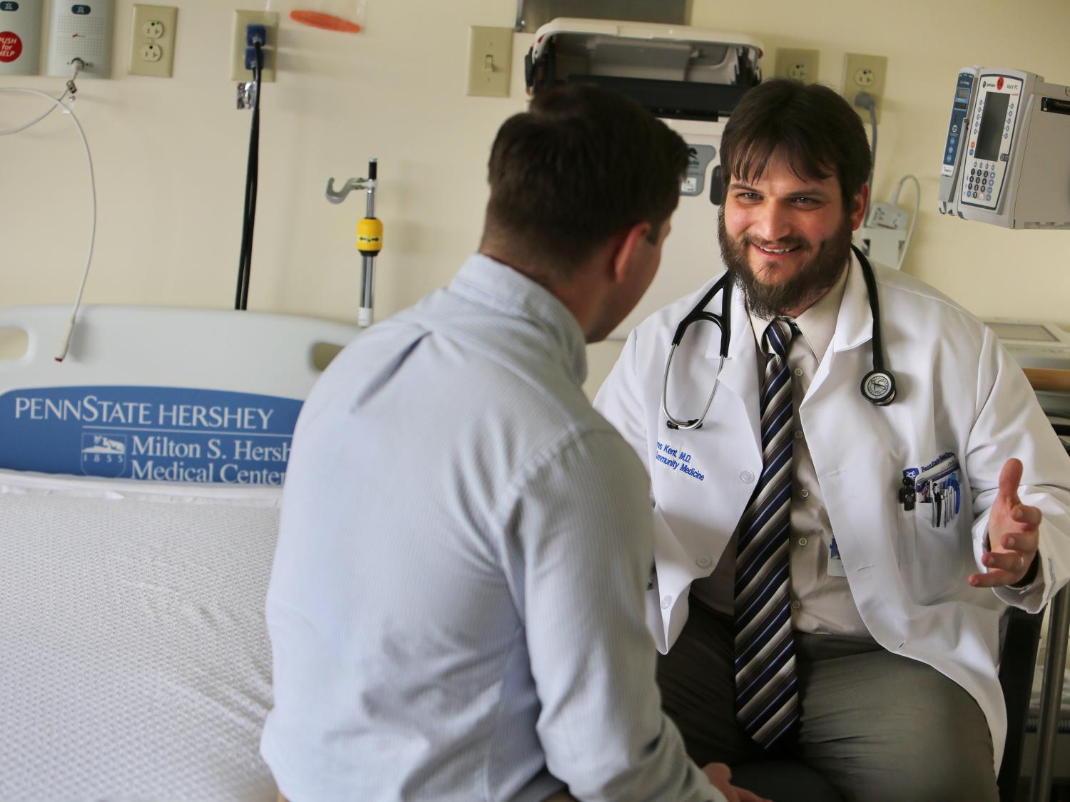 Dr. James Kent, in a white physician's coat, talks with a man in a hospital patient room. Various medical equipment is on the wall in the background.
