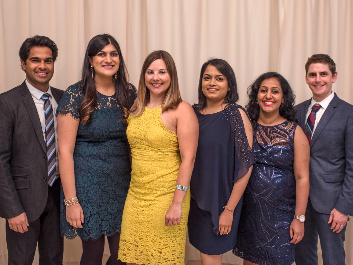 Six people -- two men and four women, wearing semi-formal attire -- stand side-by-side to pose for a photo.