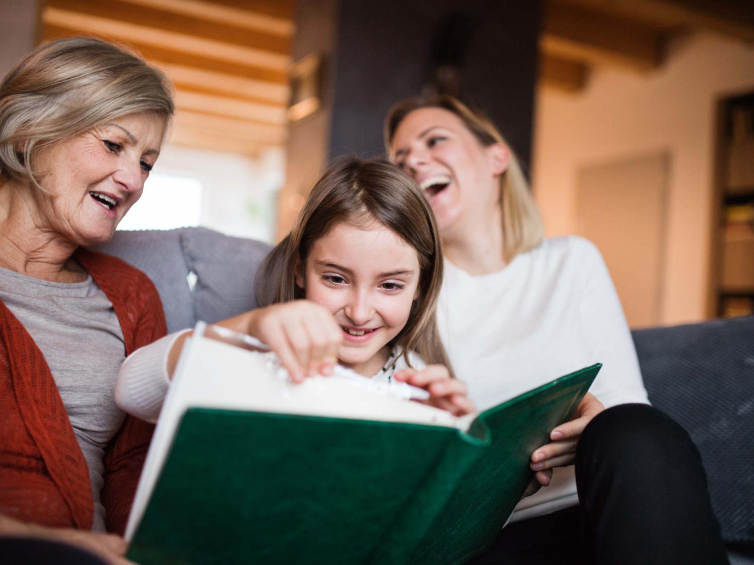 A young girl sits between two women on a couch. She holds and pages through a large photo album as the women look on, smiling. A bookshelf and window are in the background.