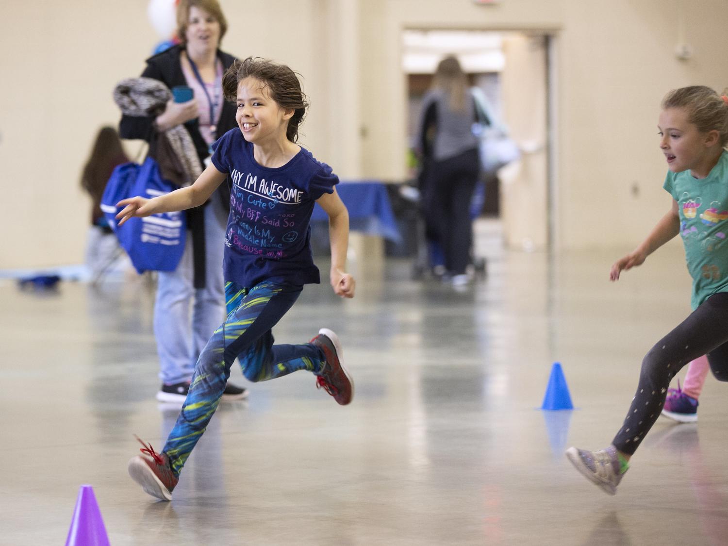 A young girl dressed in a t-shirt, long pants and sneakers smiles as she runs around a cone set up in a gymnasium. Another girl is close behind her, as a woman looks on in the background.