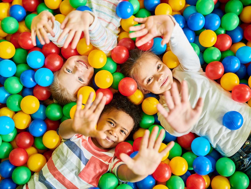An overhead view of three young children laying in a pit of colorful balls with their hands extended into the air.