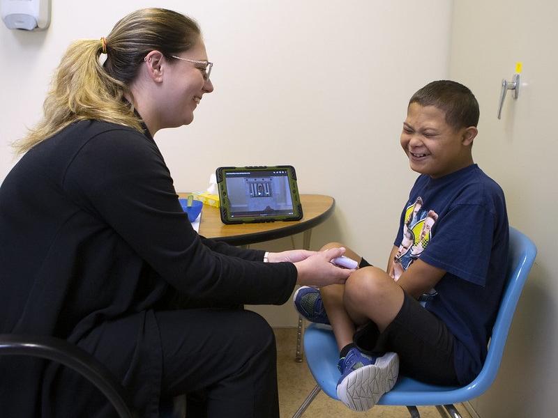 Whitney Adams, left, an intensive feeding therapist with the Penn State Children’s Hospital Feeding Program, holds out a toy to 11-year-old Jesus Rodriguez. They are both sitting in chairs at a small table. Adams is smiling. 