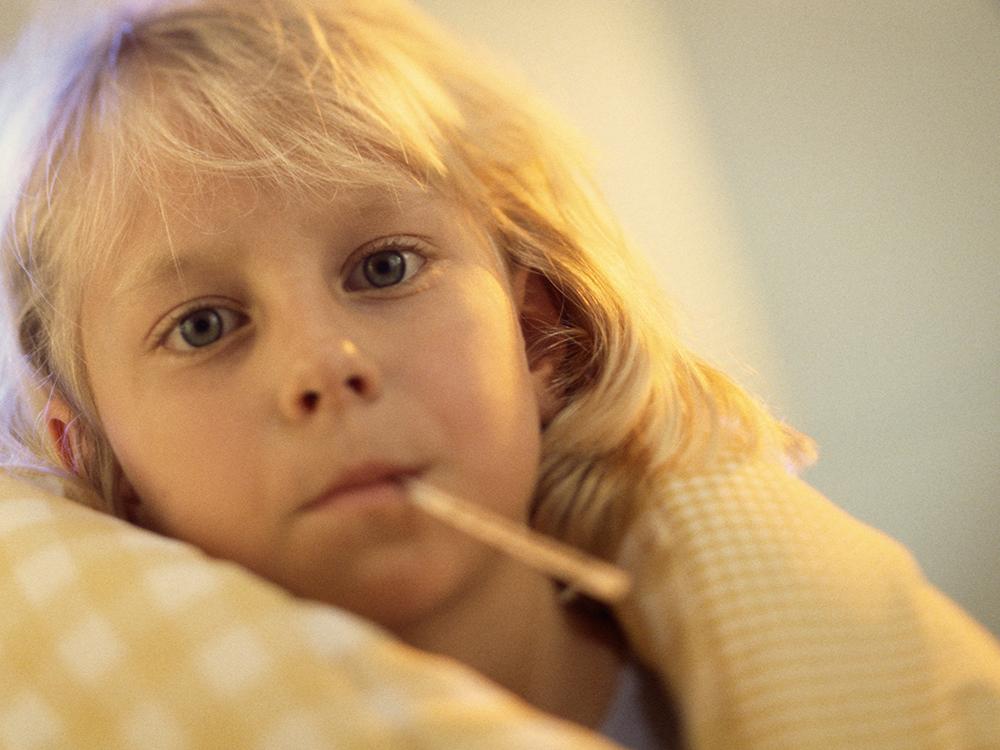 A young girl with a thermometer in her mouth is wrapped in a blanket, and looks at the camera.