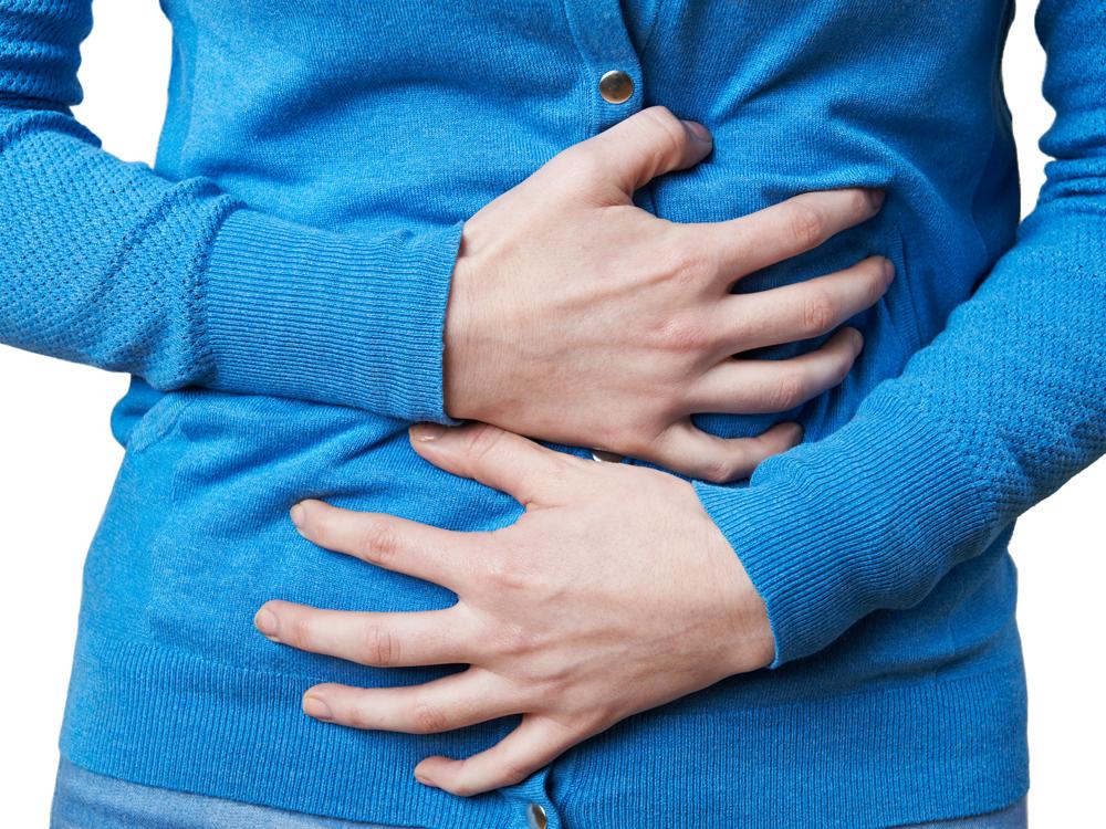 A close-up of the mid-section of a woman wearing a blue long-sleeved sweater, as she grasps her stomach and intestinal regions with both hands.