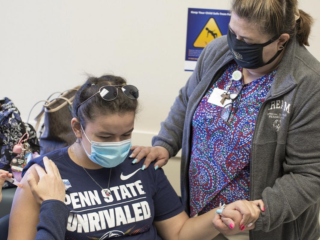 Rachel Frometa, who is seated and wears a mask and sunglasses on the top of her head, holds the hand of her mother, Betty Frometa, who stands beside her, as a vaccine is pressed into her arm by the hands of a health care worker.