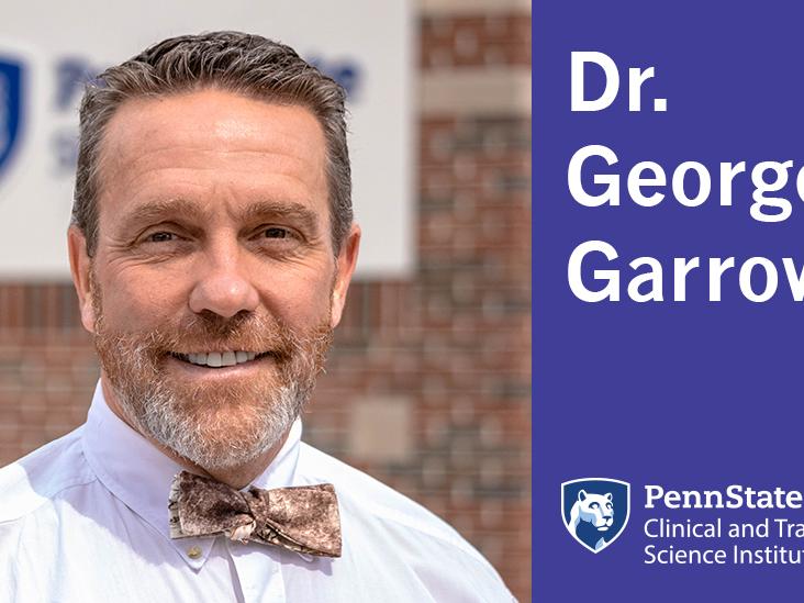 A man in a white shirt and bowtie stand in front of a Penn State sign in the background. 