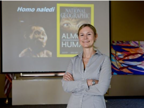 Dr. Heather Garvin poses in front of a screen, projected with imagine of Home naledi.