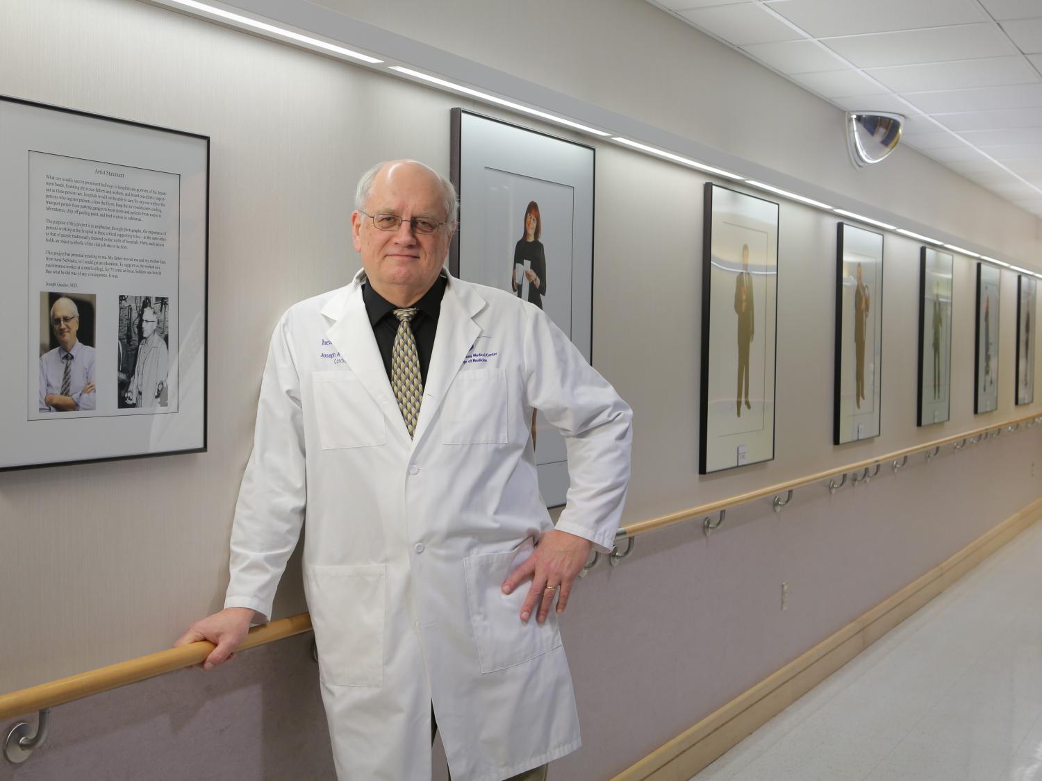 Dr. Joe Gascho wears a white coat and leans on a railing in a hallway lined with framed portrait photographs.