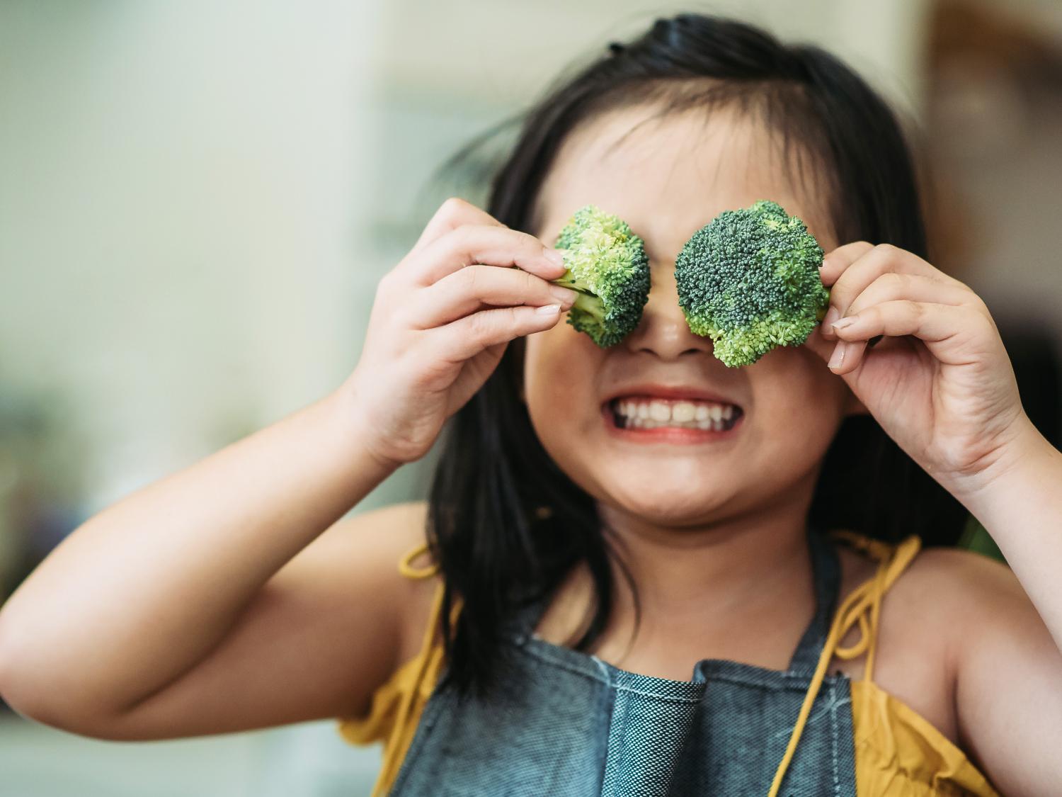 young girl holding broccoli in front of her eyes and smiling