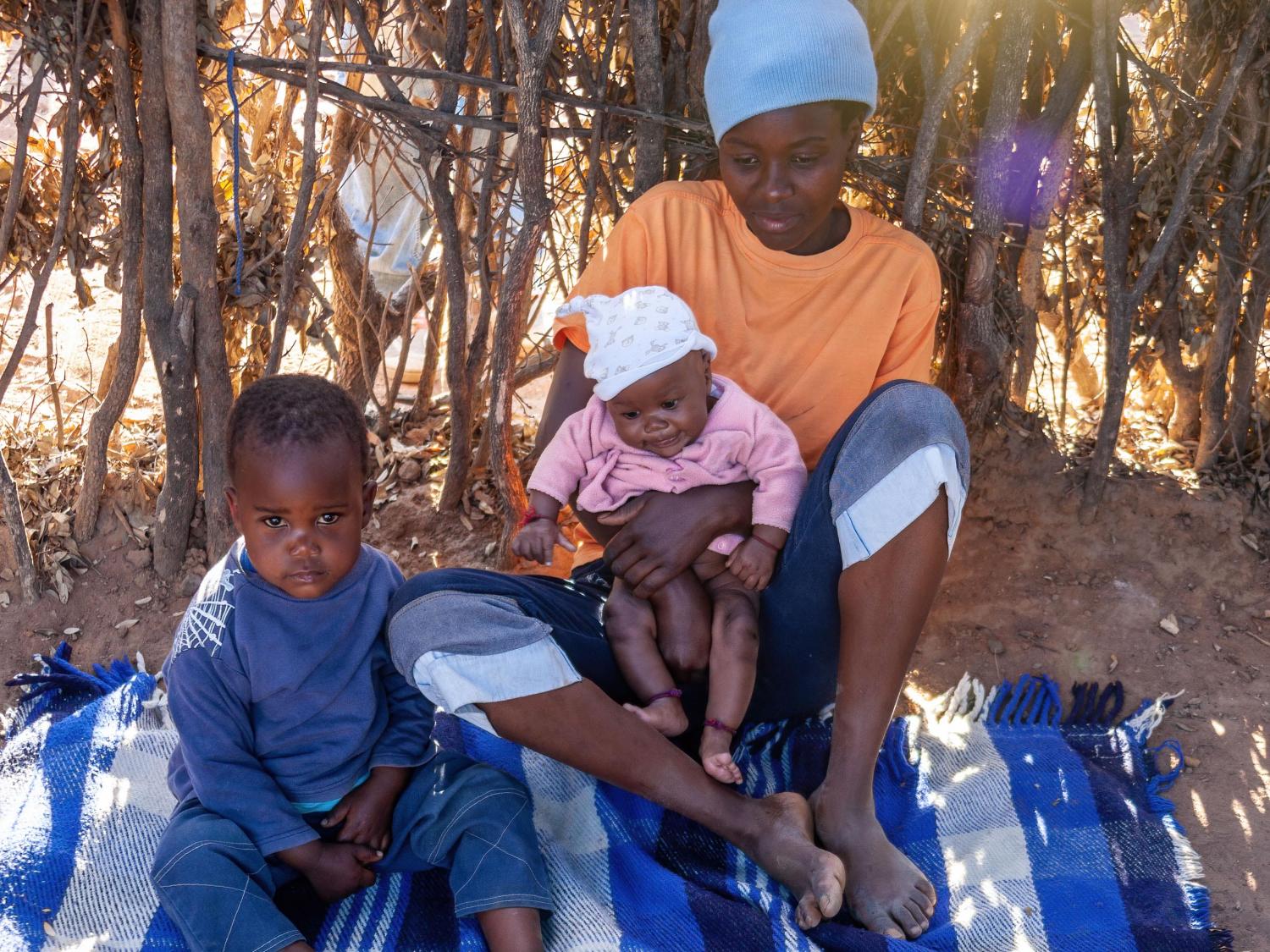 Woman sits on blanket on ground with no shoes. A small boy sits beside her, and the woman is holding a baby.