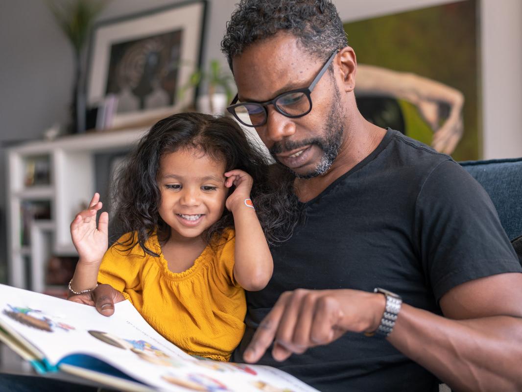 A man holds his young daughter on his lap as they both look down at a picture storybook. A bookshelf, artwork and plants are in the background.