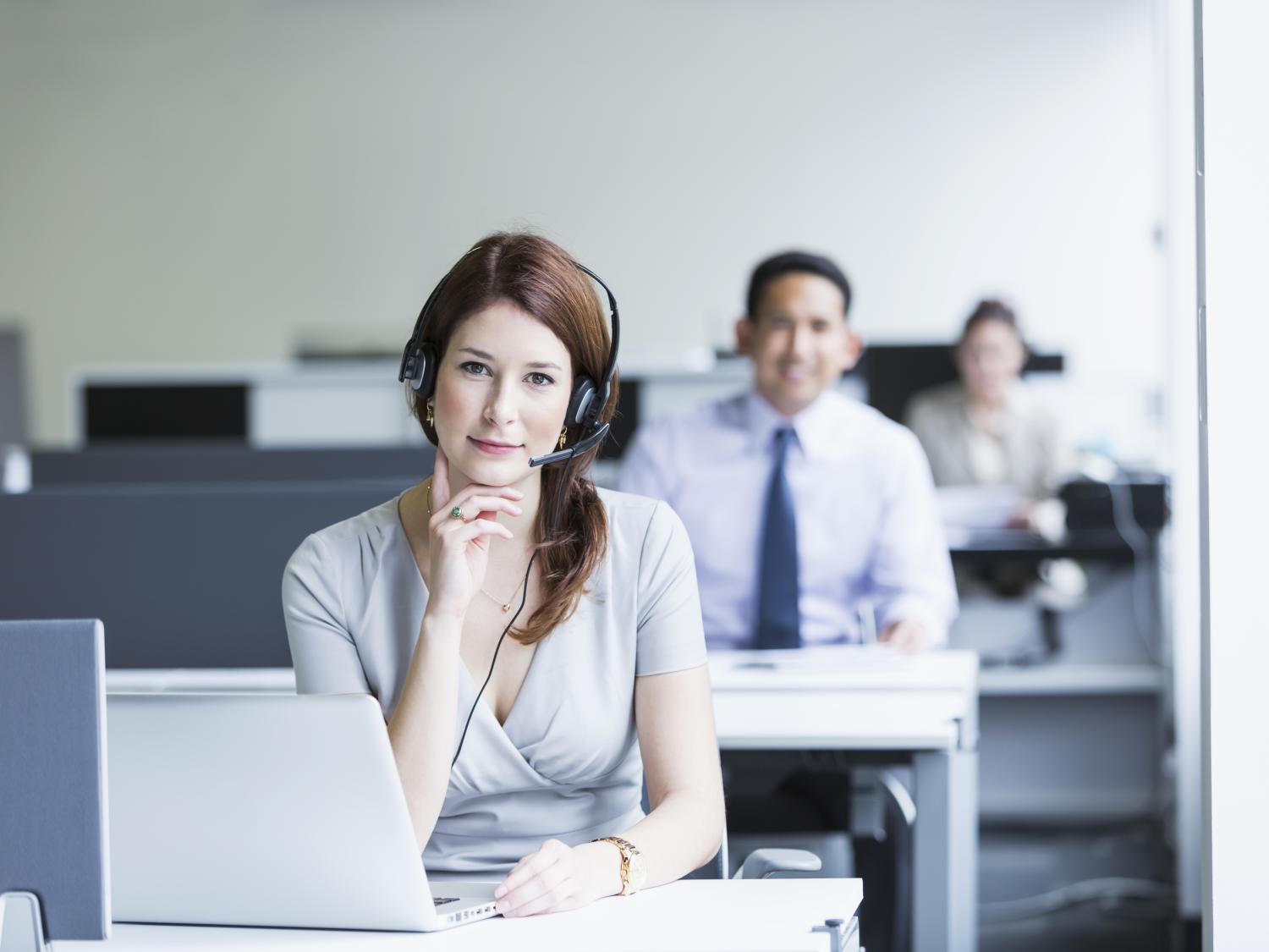 Woman in office at desk wearing headset with coworkers at desks in the background