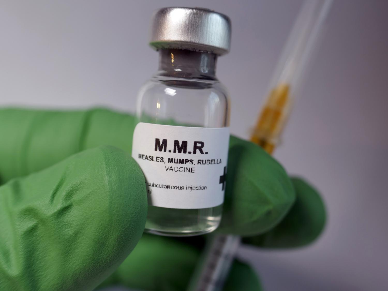 A person in a green glove holding a bottle of MMR vaccine and syringe.