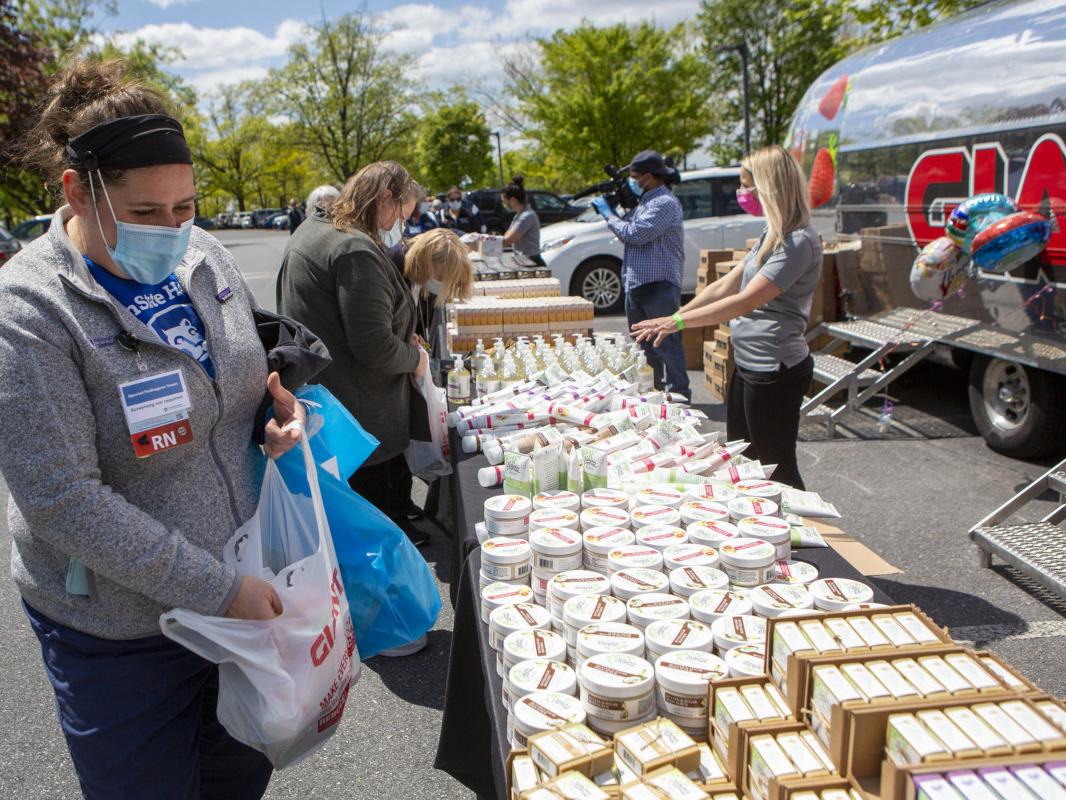 Registered nurse Lara Sowers looks over a variety of beauty care products displayed on a table set up in a parking lot. Other people are in the background, as is a silver colored travel trailer with the “GIANT” logo on it.