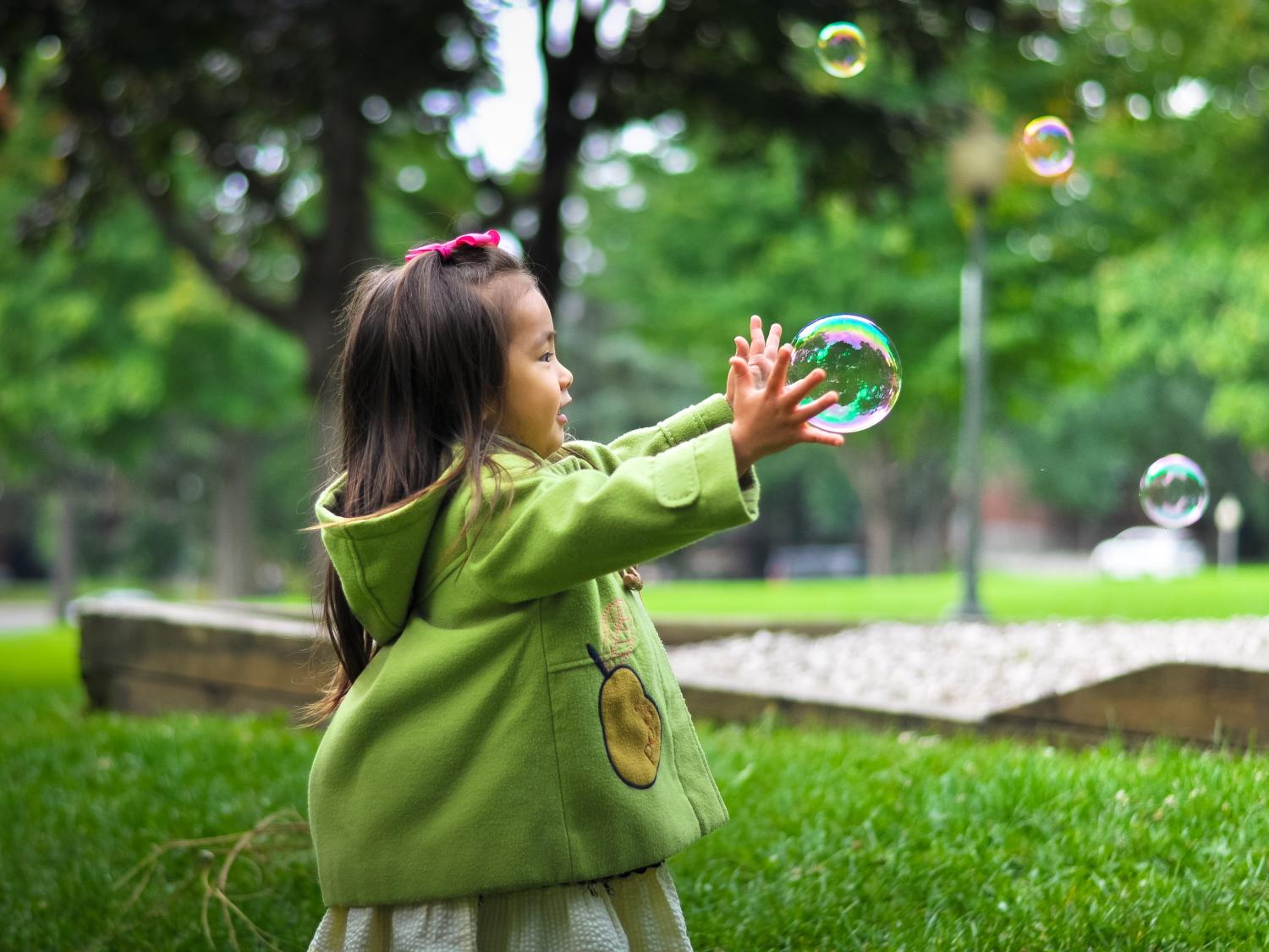 A young girl plays outside with her arms reaching to catch a bubble