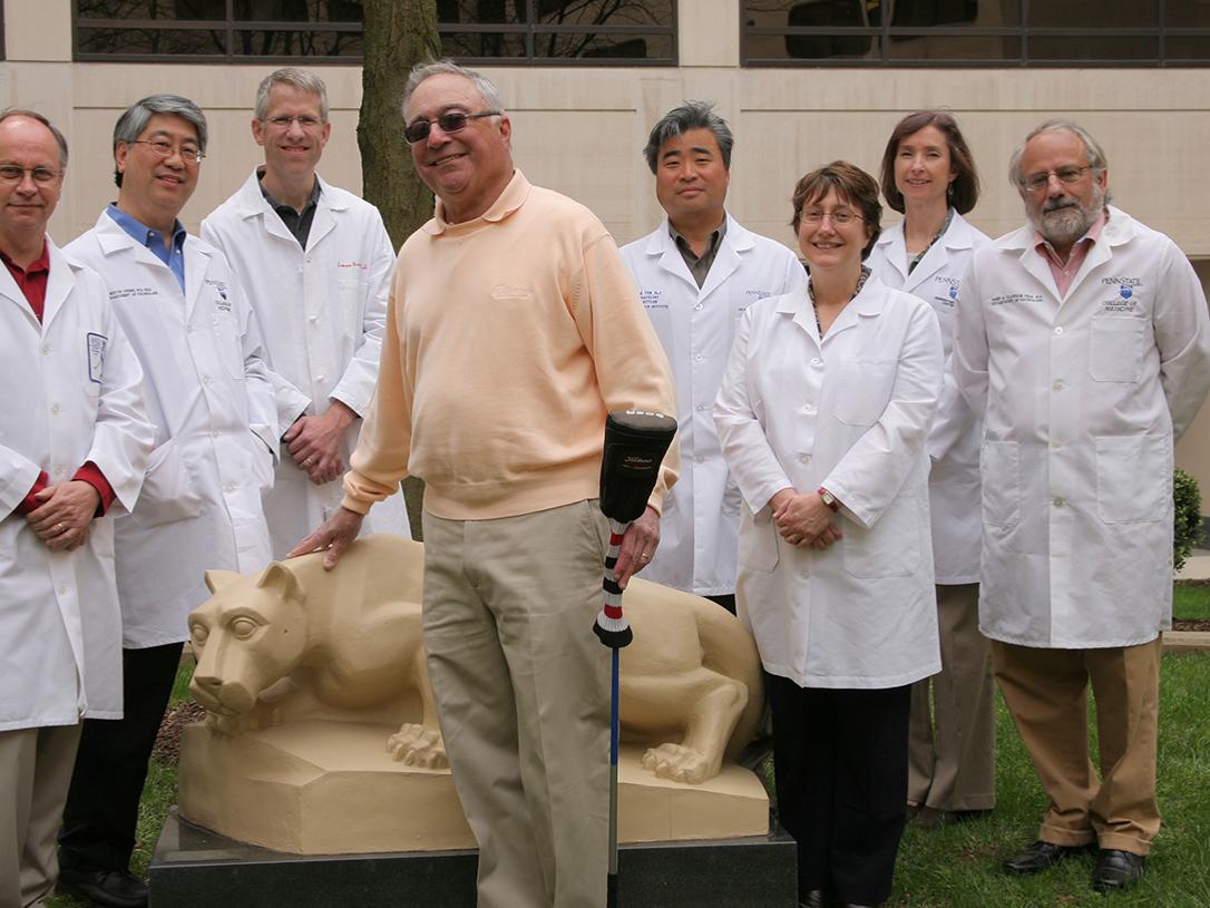 A man in a yellow sweatshirt holds a golf club and poses in front of a Nittany Lion statue, smiling. Three people pose to his left in white coats; four people pose to his right in white coats. A beige building with dark windows is in the background.
