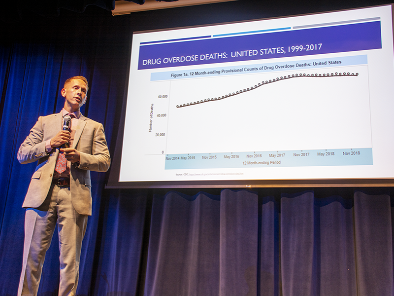 Dr. Glenn Sterner stands on stage as he delivers a speech. There is a slide behind him reviewing drug overdose deaths in the U.S. from 1999-2017 