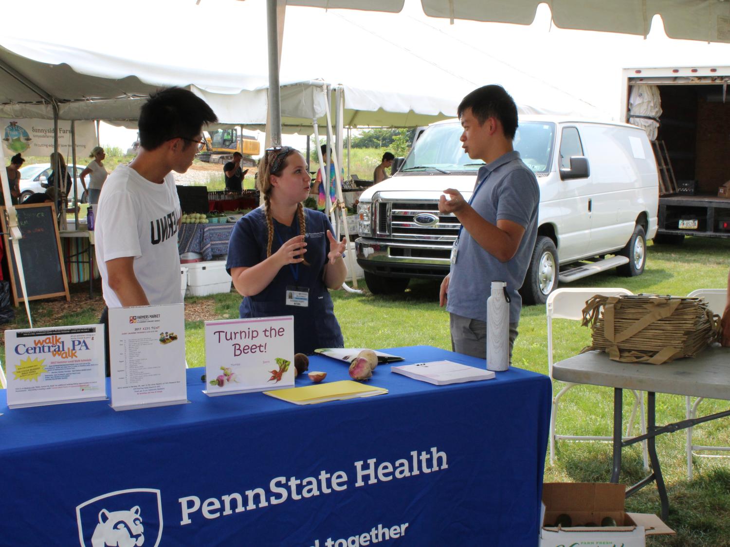 Three people – two men and one woman – stand behind a table talking with one another. The table is draped with a tablecloth with the ‘Penn State Health’ logo. It’s under a tent at an outdoor event.