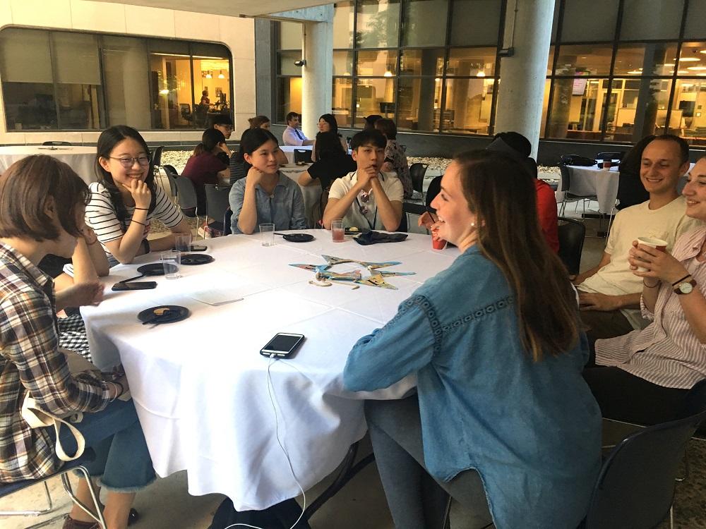 Eight international students with the 2018 Global Health Exchange Program sit at a round table in a restaurant inside Penn State College of Medicine. They are in their 20s, smiling and wearing casual clothes. Behind them people sit at other tables.