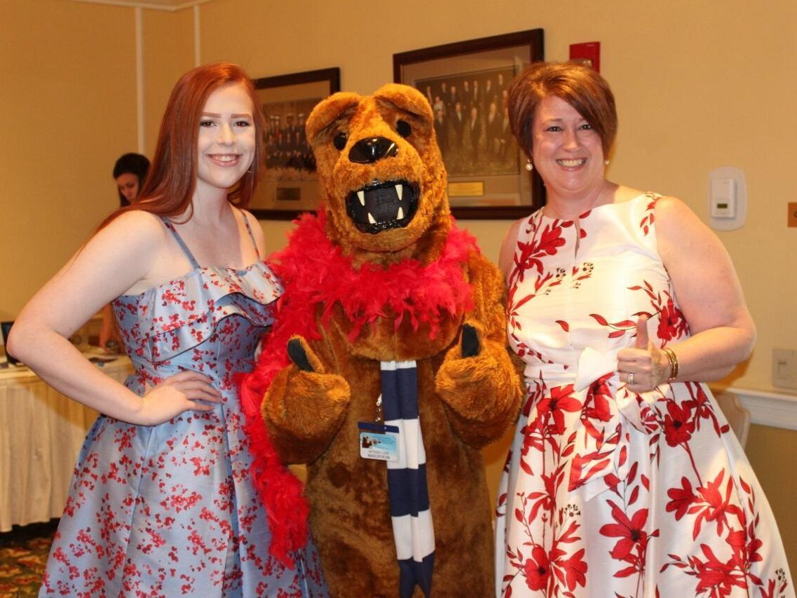 Two women in red and white dresses pose with "the Nittany Lion," who's wearing a red feather boa around its neck.