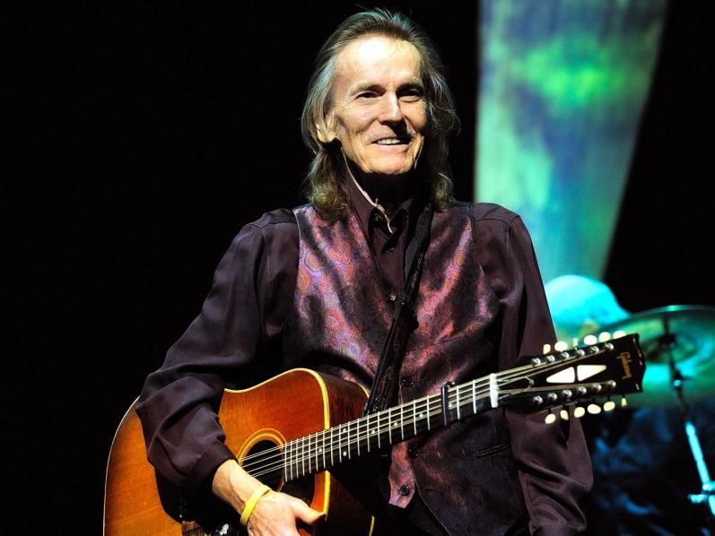 A man wearing a wearing a vest and holding an acoustic guitar smiles to an unseen audience.