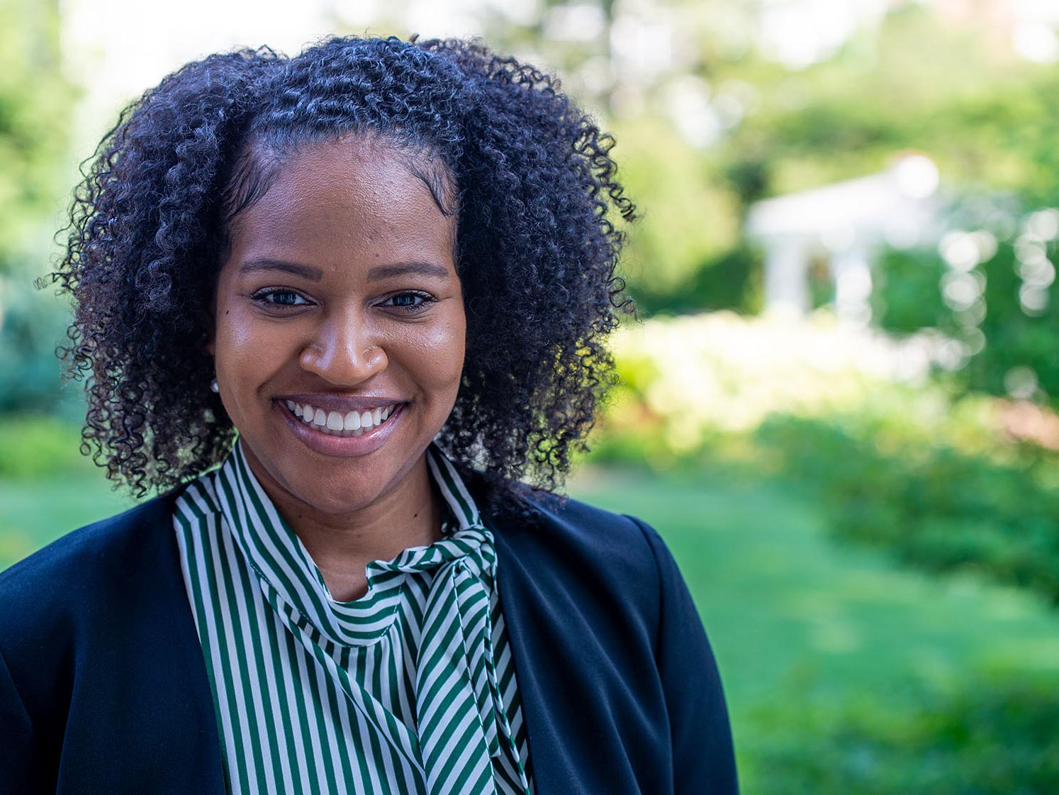 A woman faces the camera, smiling. Sunlight and greenery are behind her. 