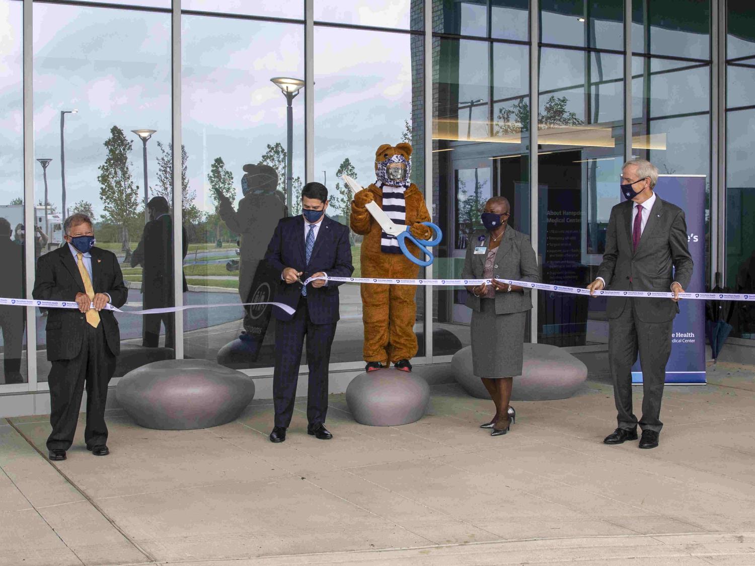A group of people and the Penn State Nittany Lion mascot stand in a line, socially distanced, holding onto a white ribbon with the Penn State Health logo on it. The mascot is positioned in the middle, holding an oversized pair of scissors.
