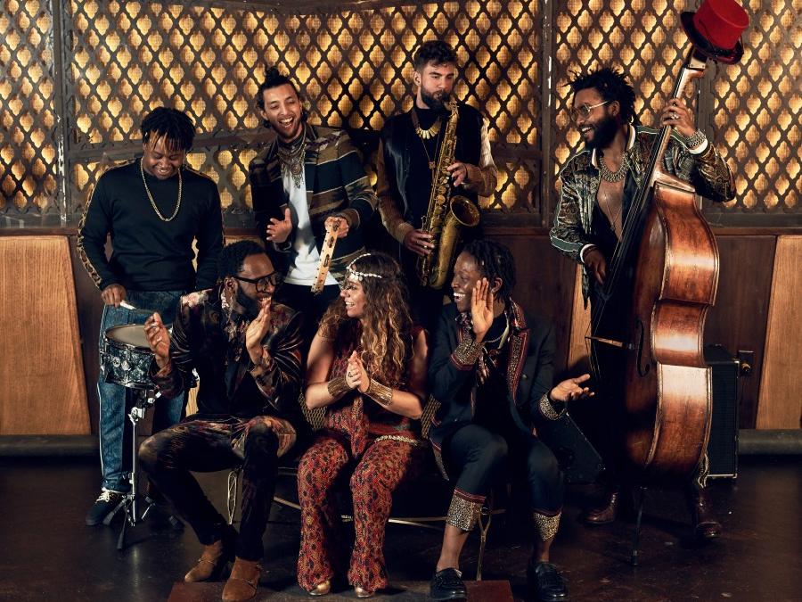 A group of musicians sit around a lounge chair and play jazz instruments and clap.