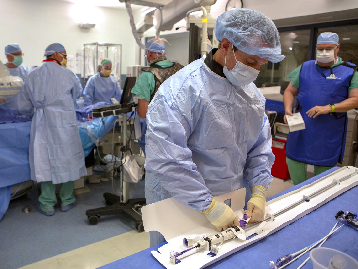 A team of six people from Penn State Heart and Vascular Institute surgeon are in an operating room and preparing for a TAVR procedure. Each are wearing surgical masks, caps and gowns. The man in the foreground is touching a heart catheter device.