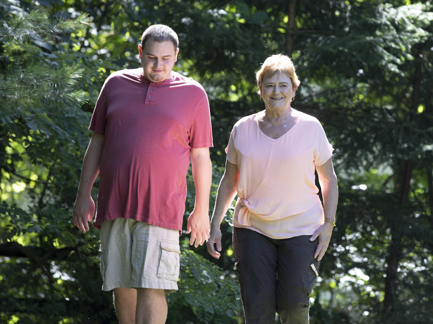 Man in red shirt and tan cargo shorts walks alongside woman in pink top and black pants in a wooded yard.