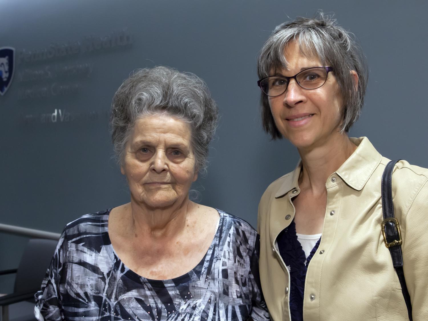 Two women stand side-by-side posing for a photo. The backdrop is a dark blue wall. A Penn State Health logo is on the wall to the left, slightly out of focus.