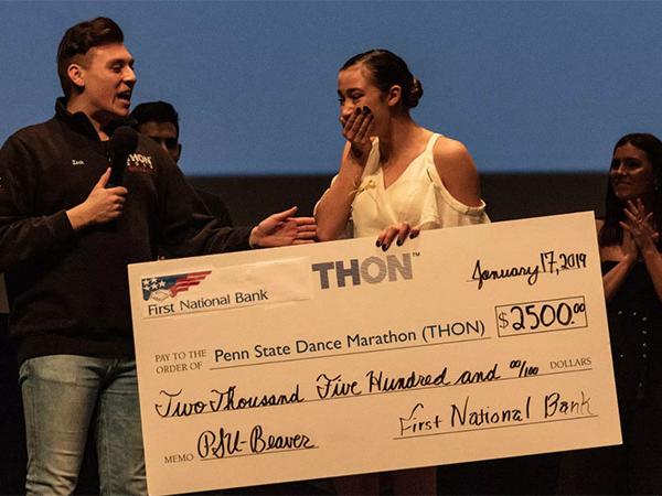 Dancer Hailey Burbage stands on stage, covers her mouth with her hand and reacts with emotion as she is presented a large check for winning the THON Showcase.