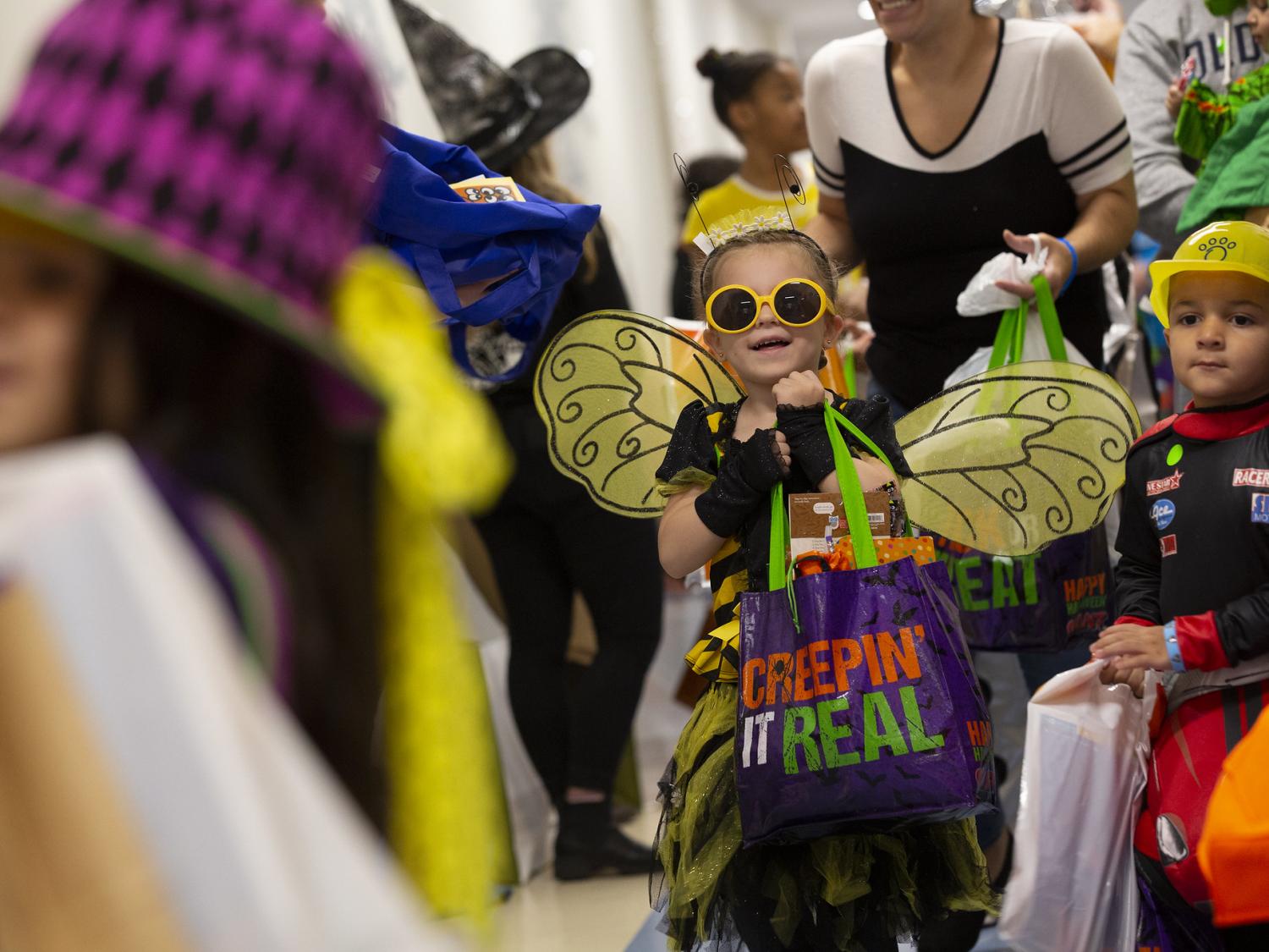 Trinity Stryker hoists a decorative bag. She’s wearing sunglasses, wings and antennae. All around her are other children and adults in costume holding bags.