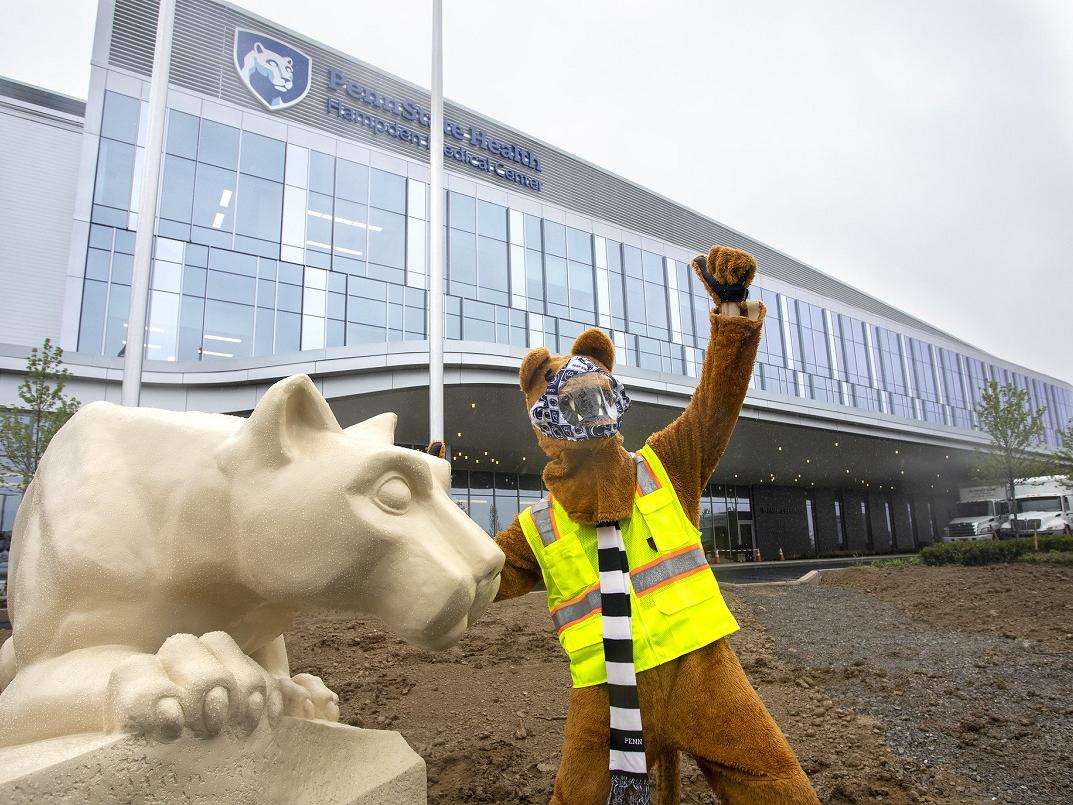 The Nittany Lion mascot raises its left arm as it touches the Nittany Lion statue with its left arm. The mascot is wearing a face mask, safety vest and scarf. Behind them is the newly constructed Penn State Health Hampden Medical Center.