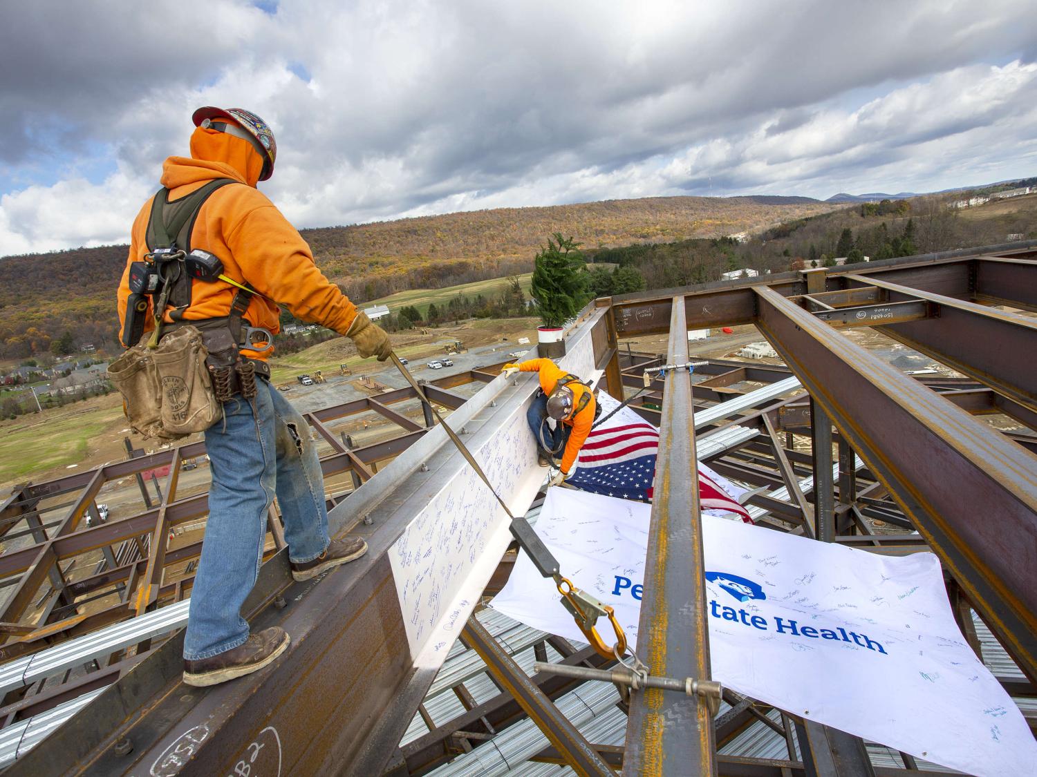 Two construction workers guide a beam into place on top of a steel structure