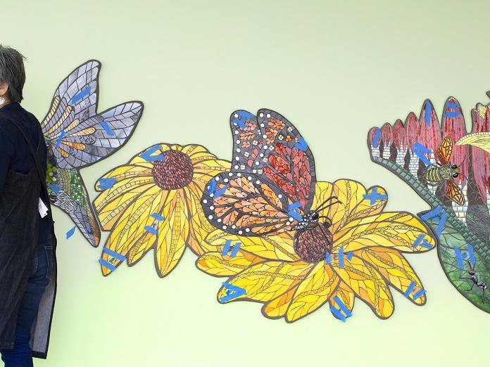 A woman in an apron glances over her shoulder in front of an image of flowers on a wall.