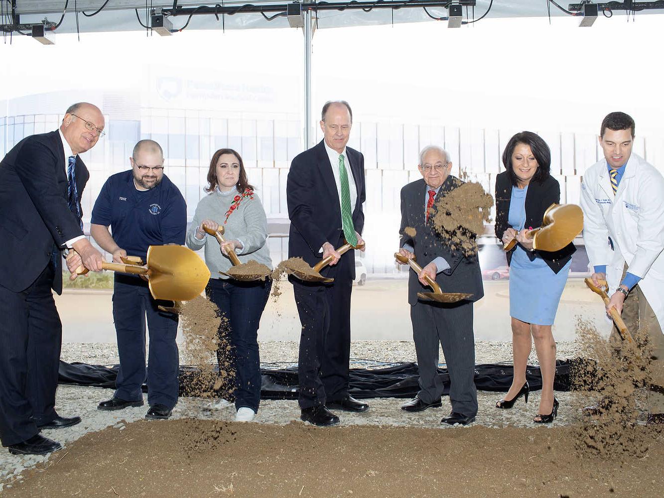 Seven people in formal attire hold gold shovels and toss dirt in the air. A large rendering of Penn State Health Hampden Medical Center is in the near background.