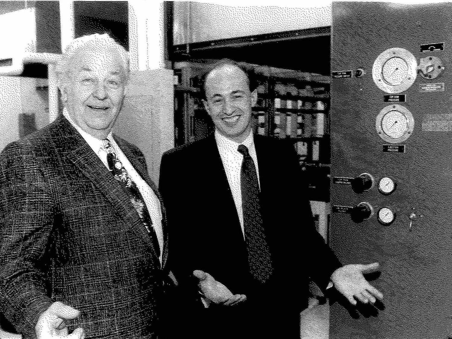 Old black and white photo of two men standing and smiling in front of lab equipment 