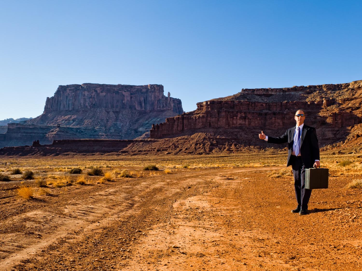 A person in a business suit trying to hitch hike in the middle of the desert