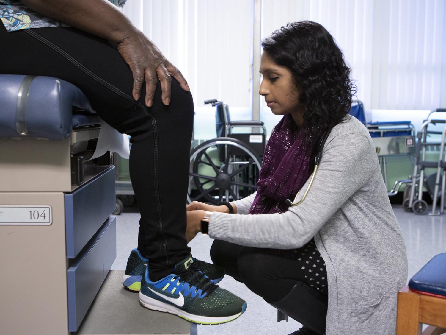 Dr. Leesha Helm crouches down to examine the legs of patient Clotilde Ngono Bodoy, who is sitting on an examination table at Beacon Clinic. A wheelchair and other medical equipment are in the background.