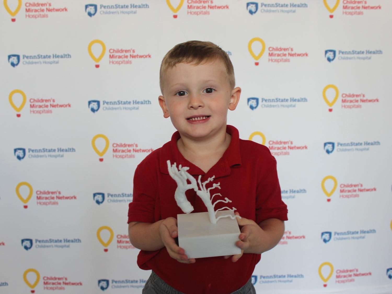 A young boy poses for a photo holding a 3-D printed model of his own heart. A step-and-repeat backdrop bears the logos of Children’s Miracle Network Hospitals and Penn State Children’s Hospital.