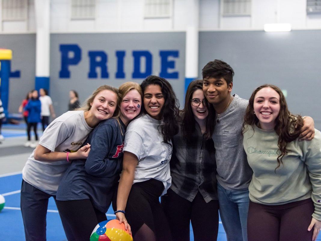 Six high school students – five female and one male – pose for a group photo with arms around each other. They’re in a school gym, where a wall with the word “PRIDE” is in the background.
