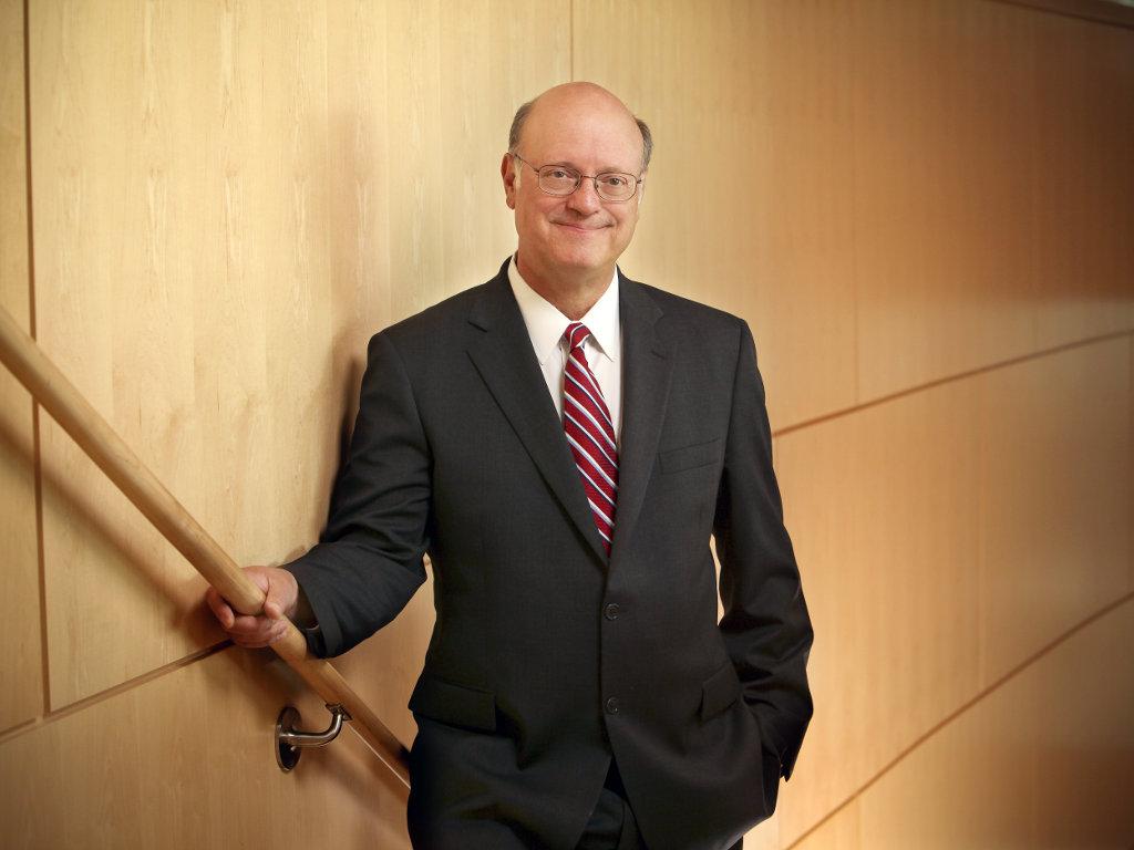 Dr. A. Craig Hillemeier poses in a dark suit with a red tie. His right hand is resting on a staircase bannister. The background is a wooden wall.