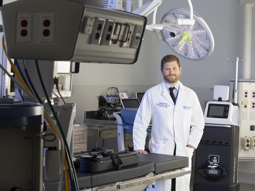 Dr. J. Nathaniel Ruhala, wearing a white medical coat, poses for a photo while standing in a hospital operating room. An overhead light and various surgical equipment is placed throughout the room.