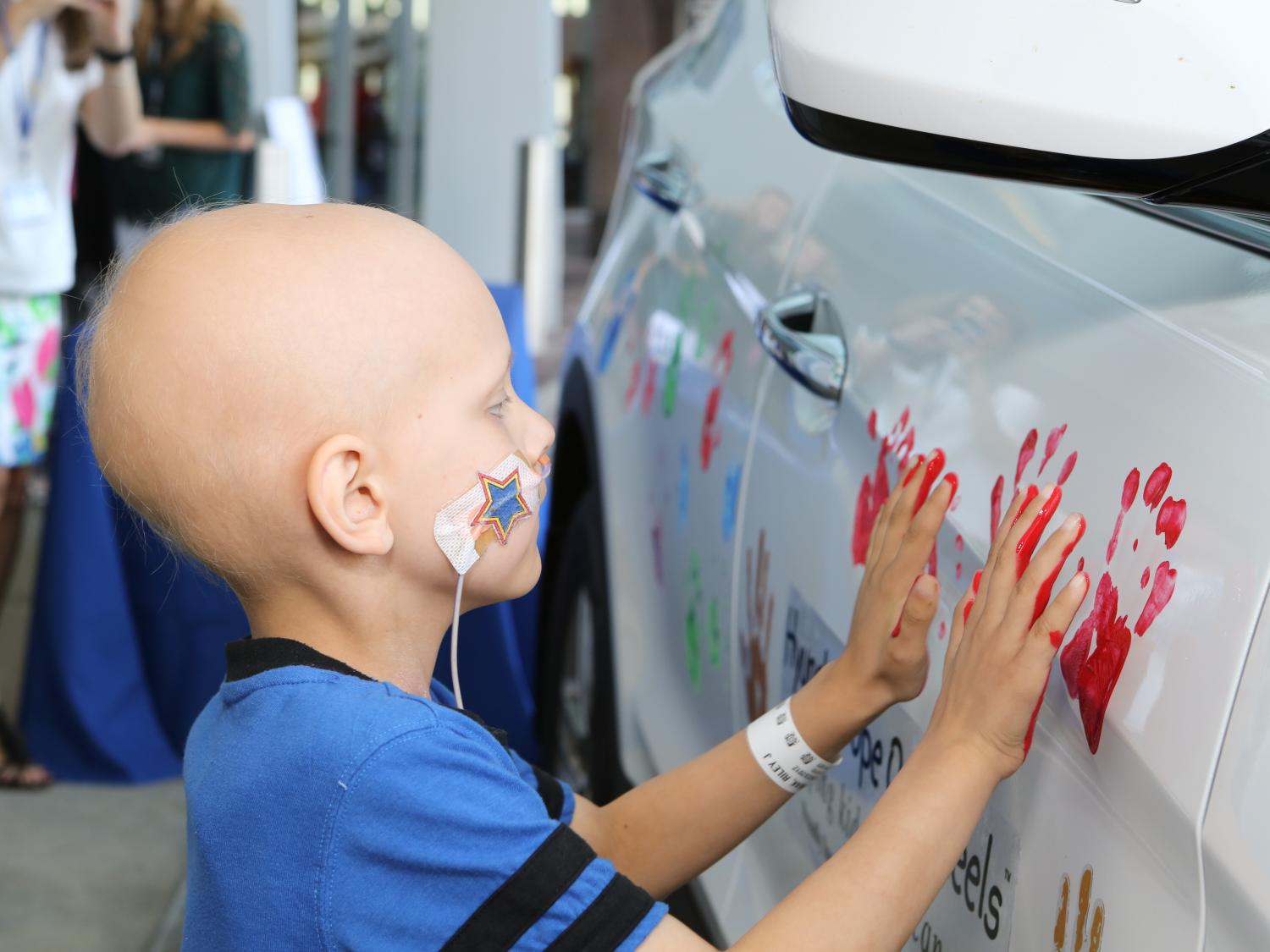 A young child in a blue shirt, and with a hospital bracelet and nasal feeding tube, places both hands – which are coated in red paint – on the side of a white car.