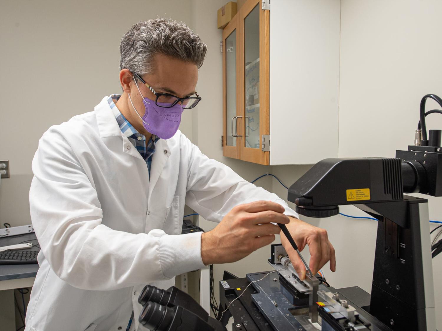 A man wearing a purple face mask adjusts a microscope with a small tool on top of a lab desk. 