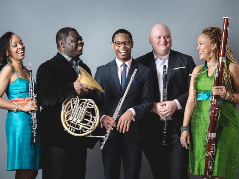 Five musicians stand and smile as they hold their instruments.